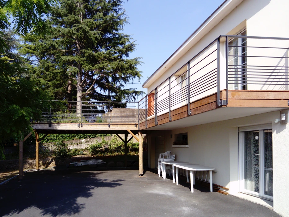 Terrasse en bois surélevée sur pilotis en bois lamellé-collé, prolongeant un balcon avec garde-corps métallique tubulaire horizontal