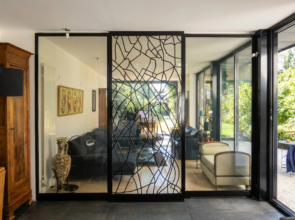 Intérieur contemporain avec cloison vitrée séparatrice en profilés acier noir, ornée d'un décor artistique en métal forgé aux motifs organiques type résille
