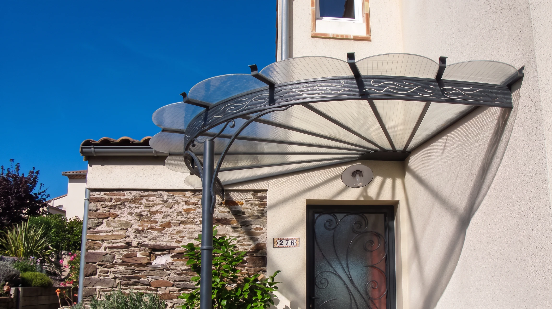 Entrée de maison numéro 276, auvent en fer forgé gris anthracite avec couverture en polycarbonate alvéolaire translucide, structure en éventail rayonnant depuis un poteau central